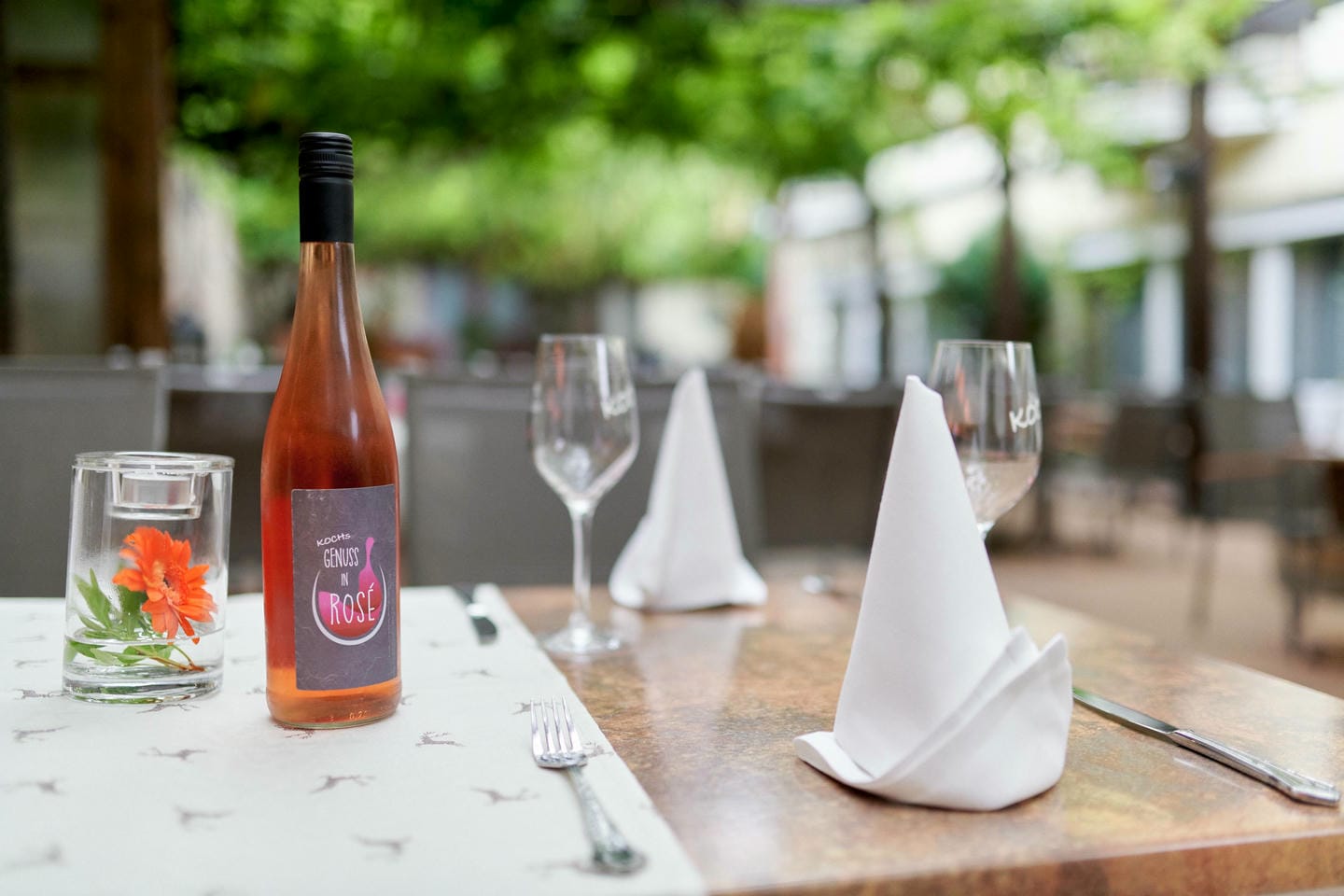 Gedeckter Tisch im Außenbereich mit einer Flasche Roséwein, dekoriert mit einem Glas mit Blume und gefalteten Stoffservietten.