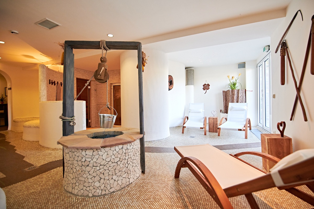 Heller Wellnessbereich mit Liegestühlen, einem Brunnen mit Eimer, Naturdeko und viel Licht.
